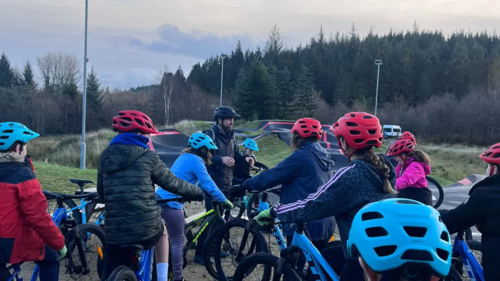 Group of children on bikes wearing helmets listening to a teacher.