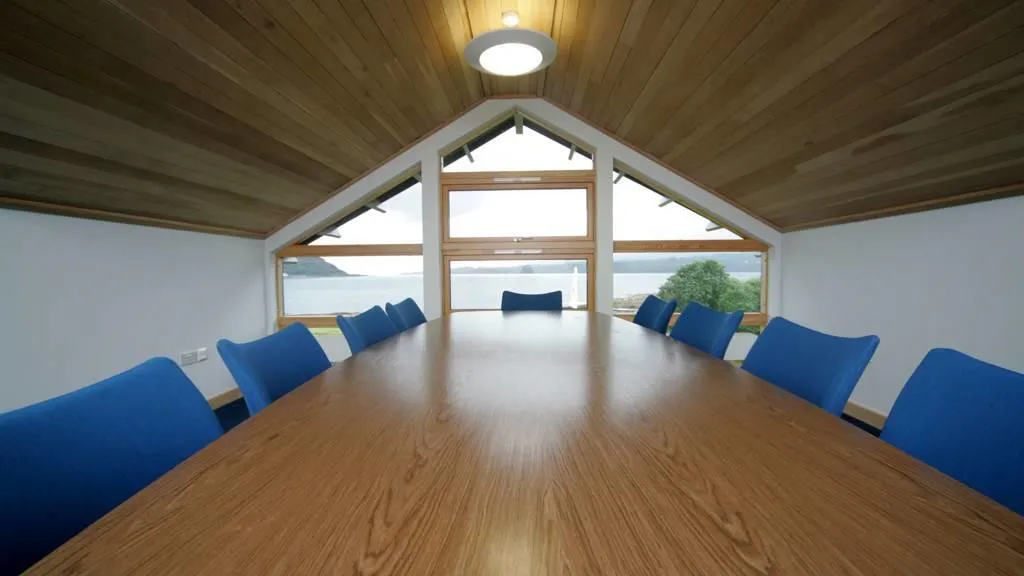 Conference room with long wooden table, surrounded by blue chairs. A large window is at the end of the room.
