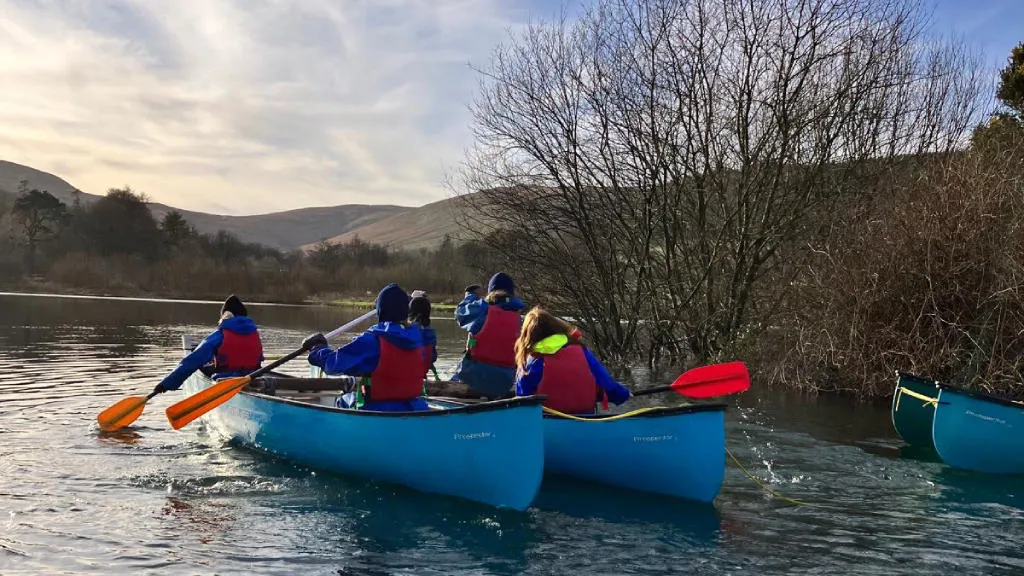 Two canoes gliding along the water with people in lifejackets steering with oars, with trees and hills in the background.