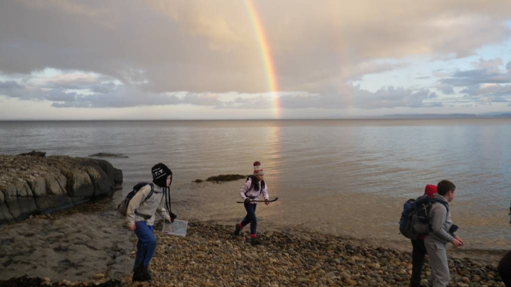 3 children on a pebble beach with a rainbow in the background