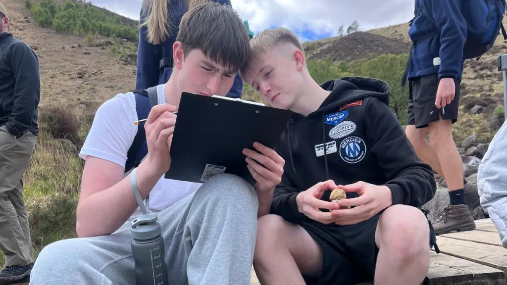 A boy writing on a notepad sitting beside his friend who is watching.